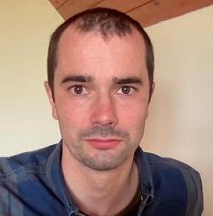 Man looking directly at the camera, with the backdrop of a magnolia coloured wall and wood coloured ceiling